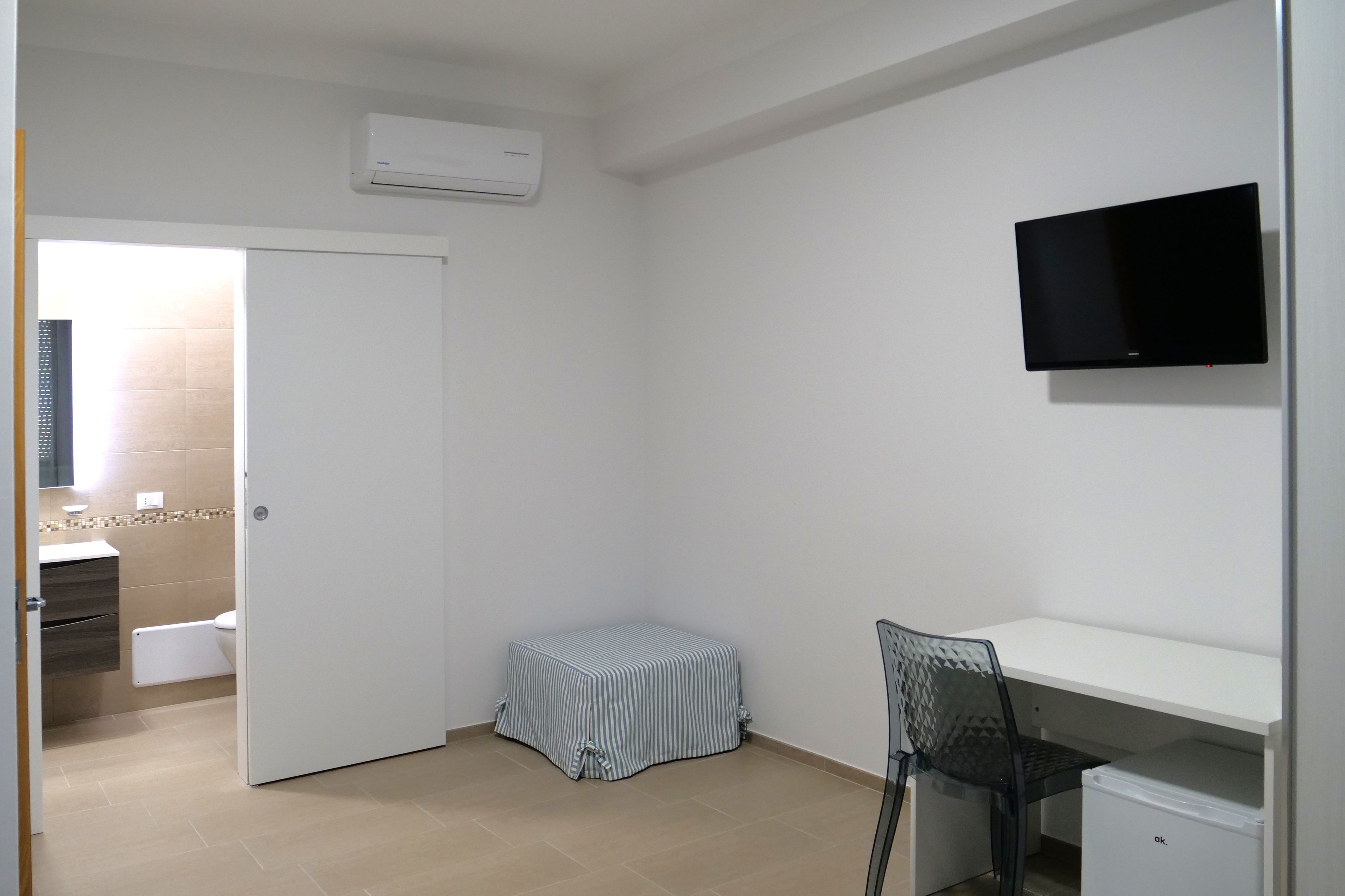 A room with a bed, a TV mounted on the wall, a small table with a chair, and a white cabinet.