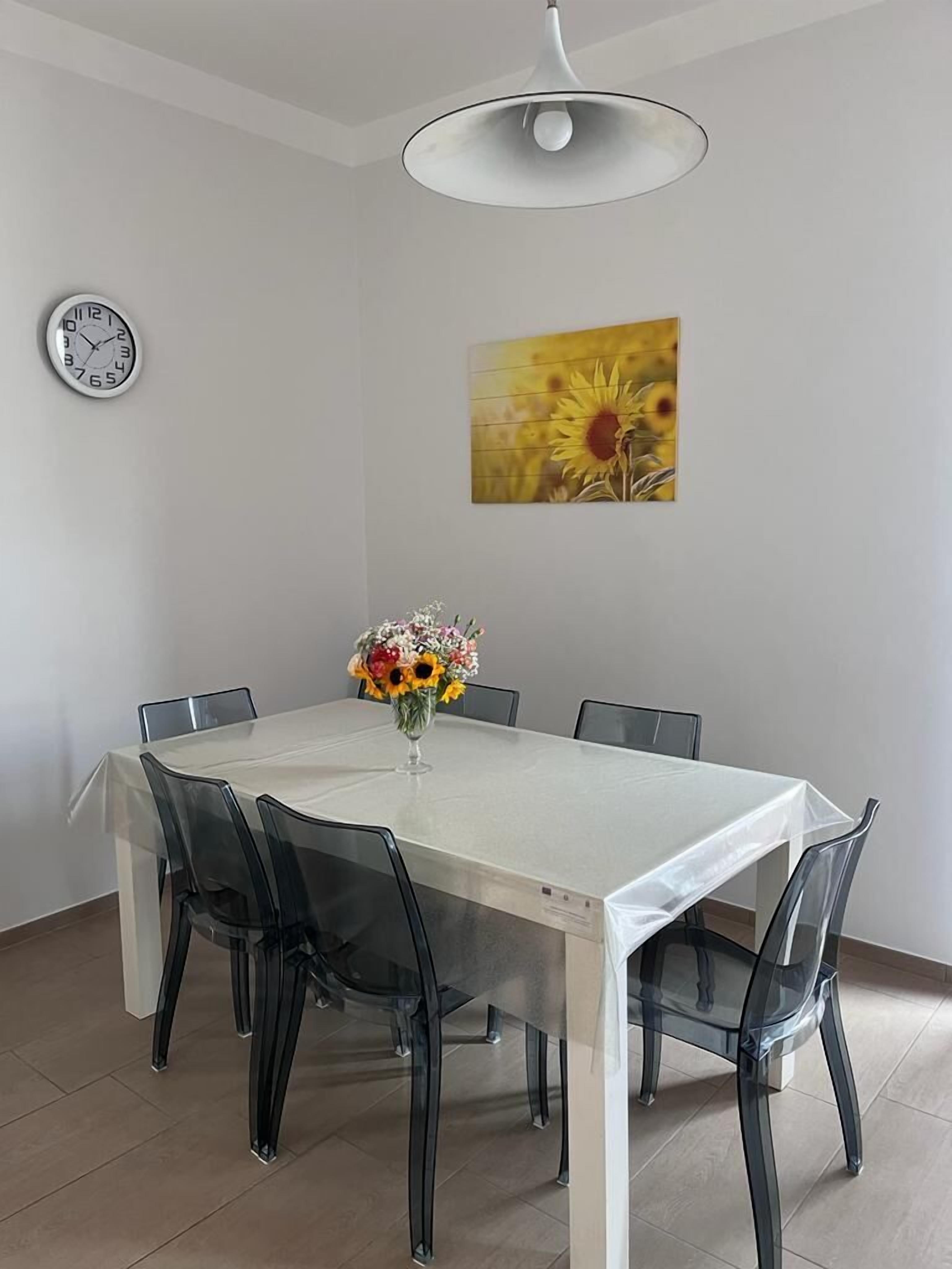 A dining area with a white table, black chairs, a clock, and a sunflower painting on the wall.