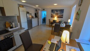 Ett kompakt kök med moderna apparater, en matplats med bord och stolar samt ett vardagsrum med en lampa och konstverk.