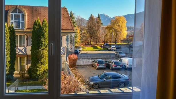 En utsikt från ett fönster med utsikt över en parkeringsplats med bilar, en byggnad med ett rött tak och berg i bakgrunden.