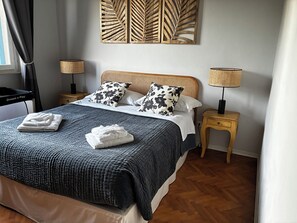 Uma cama bem arrumada com um edredom azul escuro, duas almofadas brancas e uma cabeceira de madeira. Há duas mesas de cabeceira com abajures, uma obra de arte na parede acima da cama e piso de madeira.