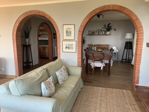 Uma sala de estar com sofá, uma mesa de jantar com cadeiras e uma cozinha visível por um arco.