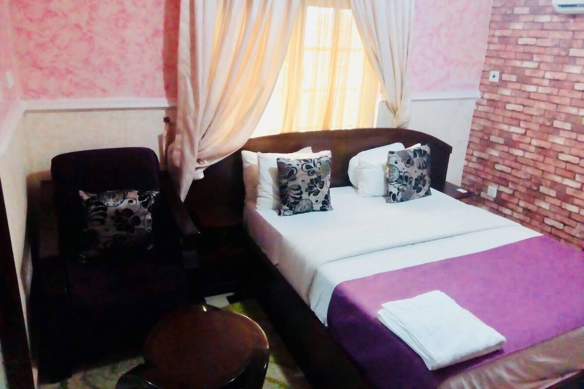 A bedroom with a bed, two pillows, a nightstand, a chair, and a window with curtains.