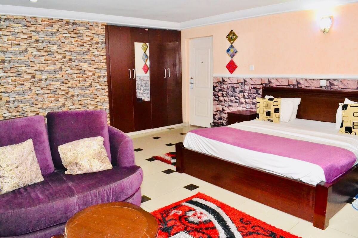 A bedroom with a bed, a purple sofa, a small round table, and a stone-patterned wall.