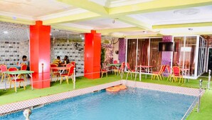 Un área de piscina interior con varias mesas y sillas, rodeada por una piscina con borde de azulejos.