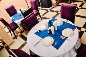 A dining area with round tables set for four, each with white tablecloths, purple chairs, and silverware. - Limoh Suites (Benin City)