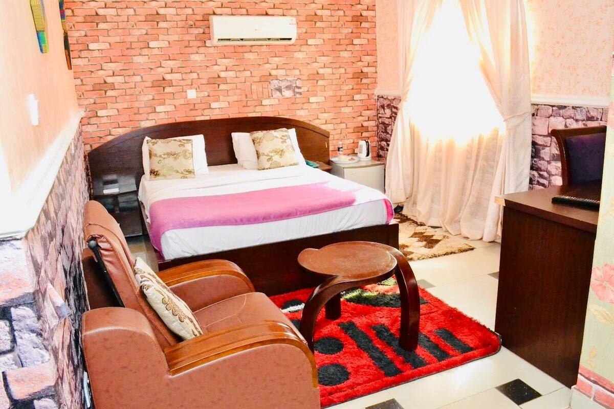 A hotel room with a brick wall, a bed with a pink bedspread, a wooden headboard, a small wooden table, a red and black rug, and a sofa with cushions.
