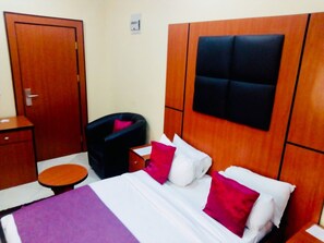 A hotel room with a wooden headboard, a bed with white and pink pillows, a small round table, and a dark armchair. - Limoh Suites (Benin City)