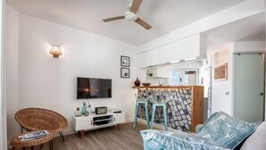 A modern living room with a flat-screen TV, wicker chairs, a sofa with patterned cushions, a kitchen area with a stone backsplash, and a ceiling fan.