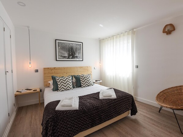 A modern bedroom with a wooden headboard, a large bed with a black and brown blanket, two pillows, a bedside table with a lamp, and a wicker chair. - Apt. Largo do Postigo (Lagos)