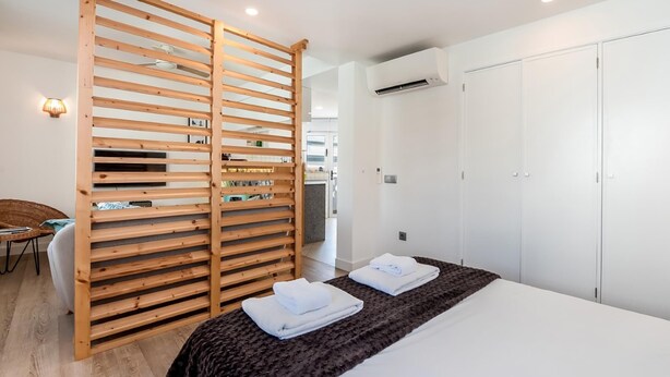 A modern hotel room with a wooden partition, a bed with white linens, a wardrobe, and an air conditioning unit.