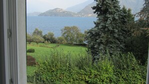 A view through a window of a grassy area with trees, a body of water, and mountains in the distance. - Situated in a beautiful & relaxing place on Lake Como,. Perfect family of 4. (Musso)