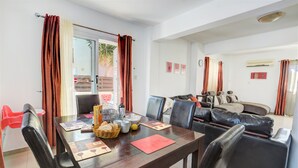 Un coin repas avec une table en bois, des chaises et un set de table rouge. On aperçoit un canapé et une fenêtre avec des rideaux au fond.