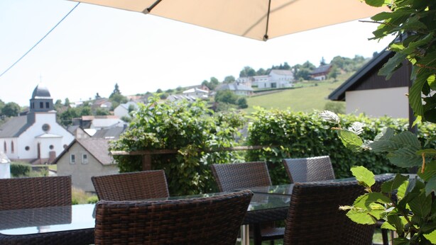 Un patio al aire libre con mobiliario de mimbre y una mesa de vidrio, con vistas a un pueblo con edificios y áreas verdes.
