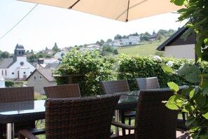 Eine Terrasss mit Korbmöbeln und einem Glastisch, mit Blick auf eine Stadt mit Gebäuden und Grünflächen.