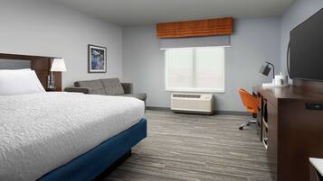 A hotel room with a large bed, a desk with a computer, an air conditioner, and a window with blinds.