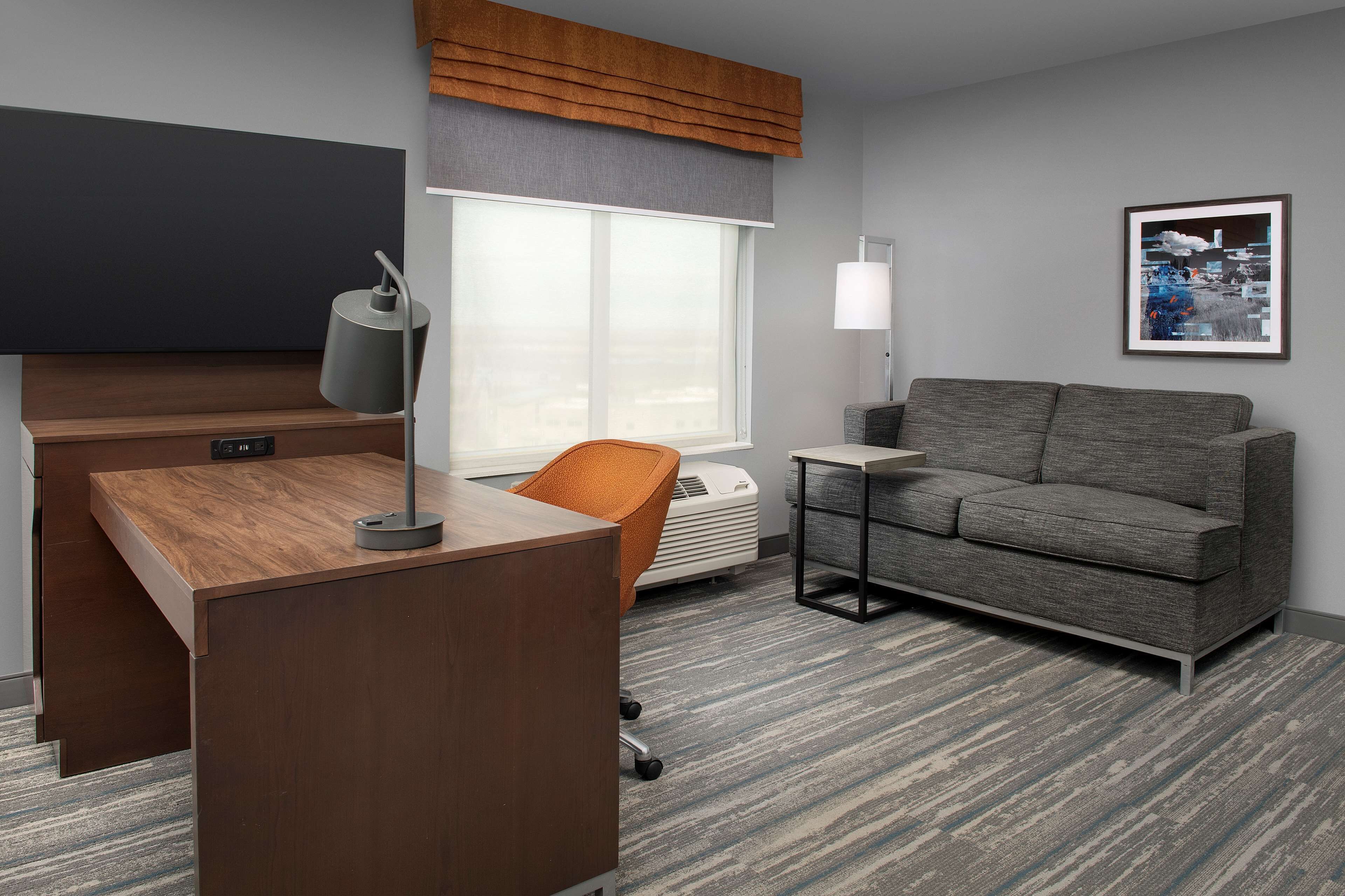 Een moderne hotelkamer met een houten bureau, een oranje bureaustoel, een grijze bank, een groot raam met jaloezieën, een staande lamp en een ingelijst schilderij aan de muur.