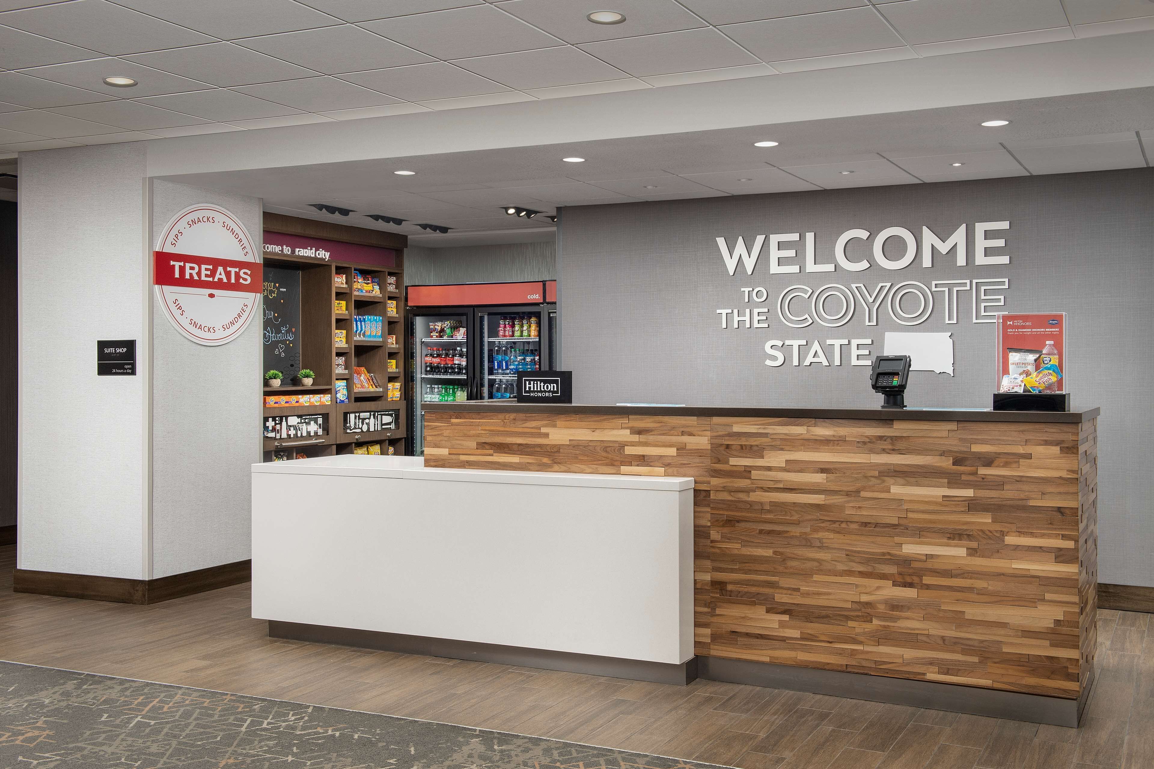 A reception desk with a 'Welcome to the Coyote State' sign, a 'Treats' sign, and a minibar area with a Hilton sign.