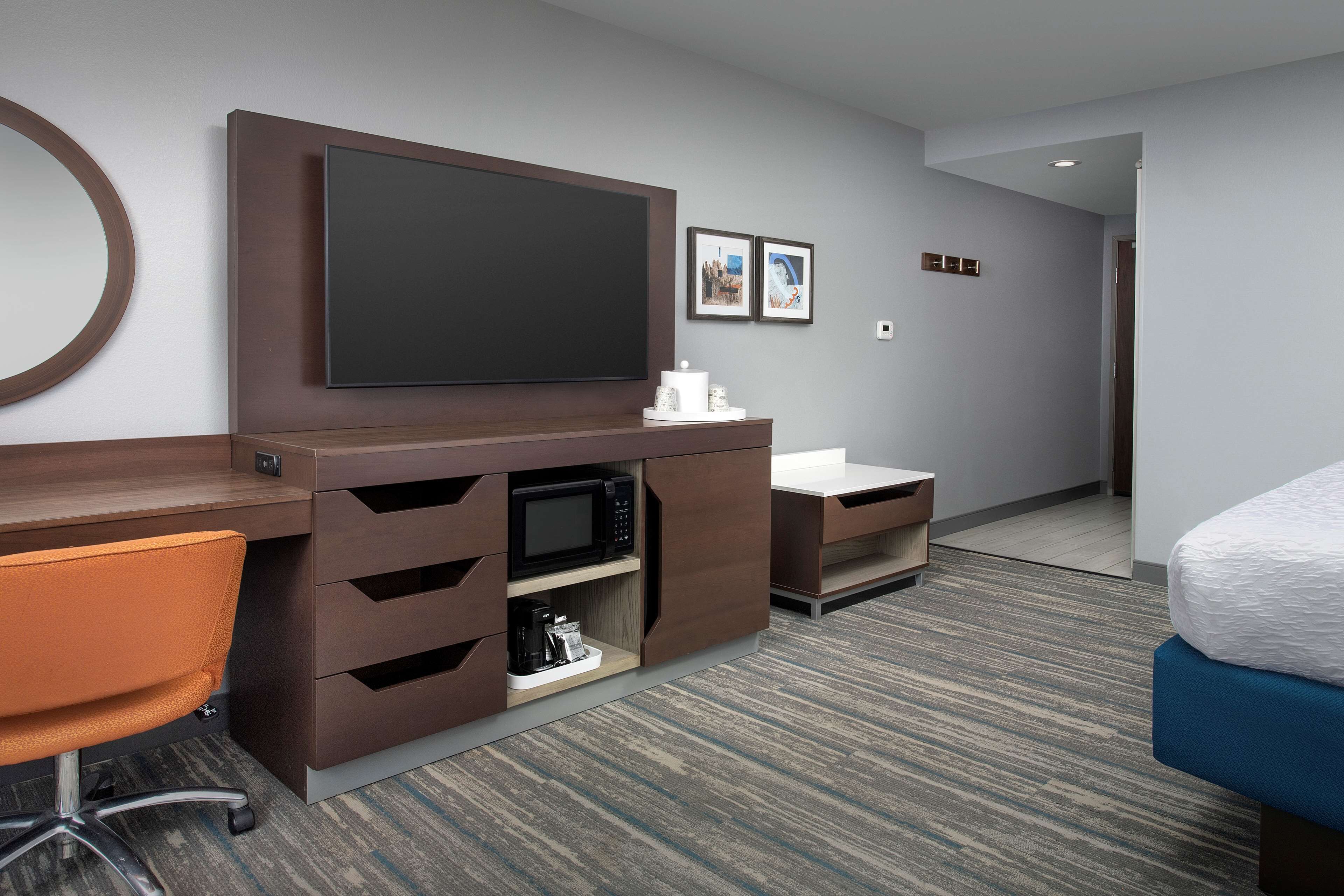 A hotel room with a large flat-screen TV, a desk with a chair, a microwave, a coffee maker, a bedside table, and a bed.
