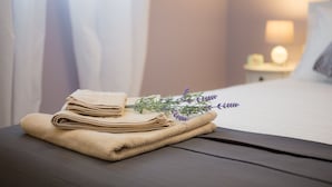 En prydligt vikt, beige handduk ligger på ett träbord bredvid sängen, tillsammans med en liten kvist lavendel.