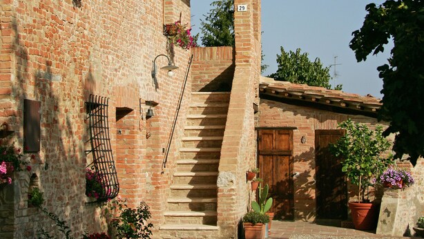 Un bâtiment en briques avec un escalier menant à une porte en bois, entouré de plantes en pot et bordé d’un chemin de gravier.
