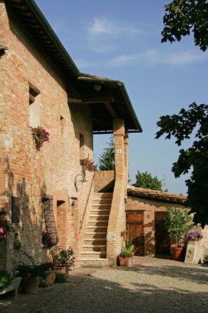 A brick building with a staircase leading to a wooden door, surrounded by potted plants and a gravel path. - Podere Il Pero (Siena)
