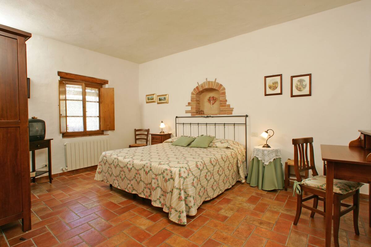 A bedroom with a bed, a wooden headboard, a window, a radiator, a table, chairs, and a television.