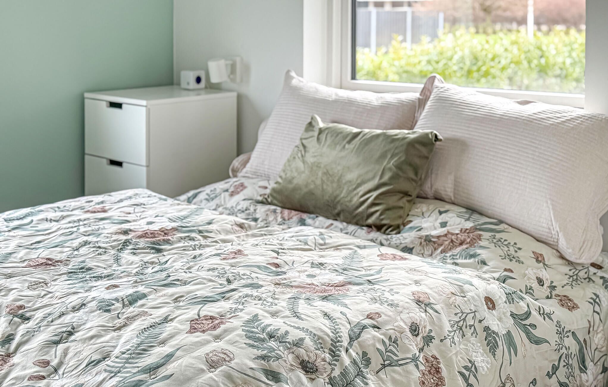 A bed with floral bedding, a nightstand with a lamp, and a window with a view of greenery.