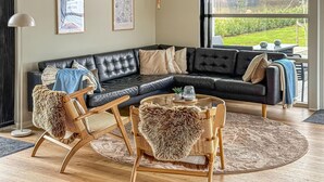 A modern living room with a sectional sofa, two wooden chairs, a coffee table, and a large window offering a view of the outdoors.