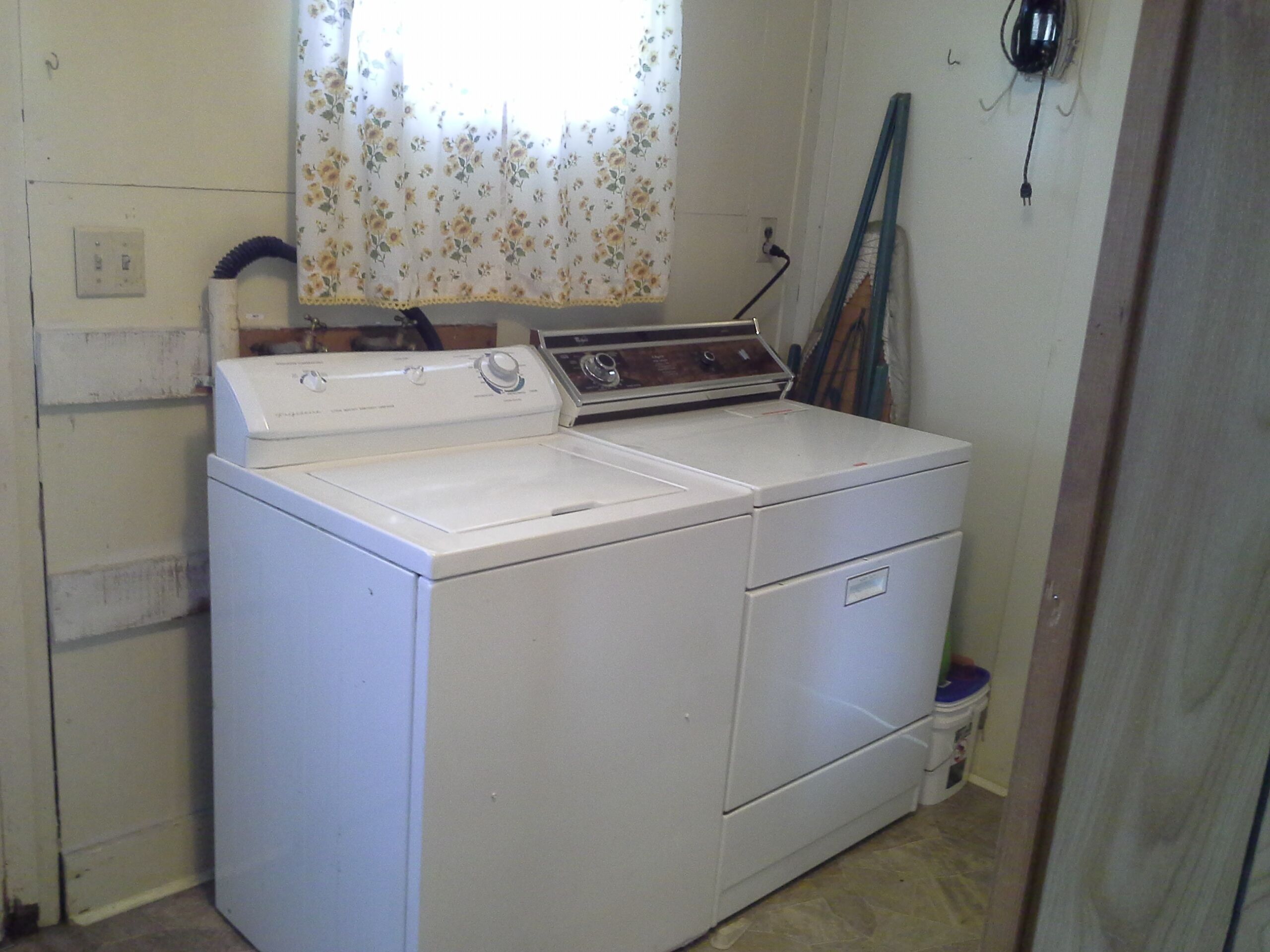 A laundry room with a front-loading washer and dryer, a window with floral curtains, and a wall-mounted electrical outlet.