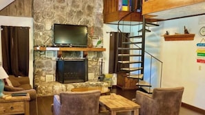 A cozy living room with a stone wall, a fireplace, a television, and a wooden staircase. - Fantastic Pool, Hot Tub, Cabana, Mt. Views, Party Pavilion, 2 Minutes 2 Frio! (Concan)