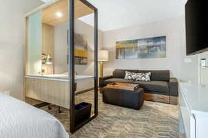 A hotel room with a bed, a sofa, a desk with a lamp, a mirror, and artwork on the wall.