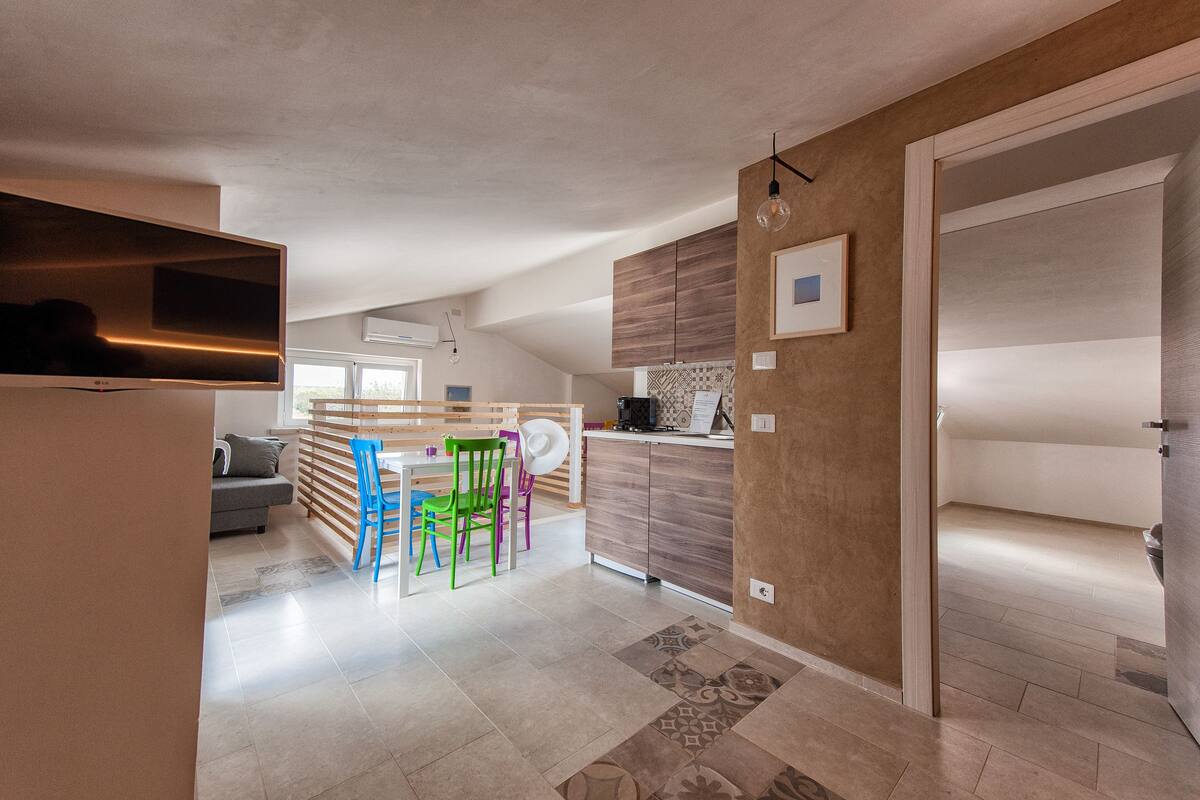 A modern kitchen with wooden cabinets, a dining area with colorful chairs, and a large window providing natural light.