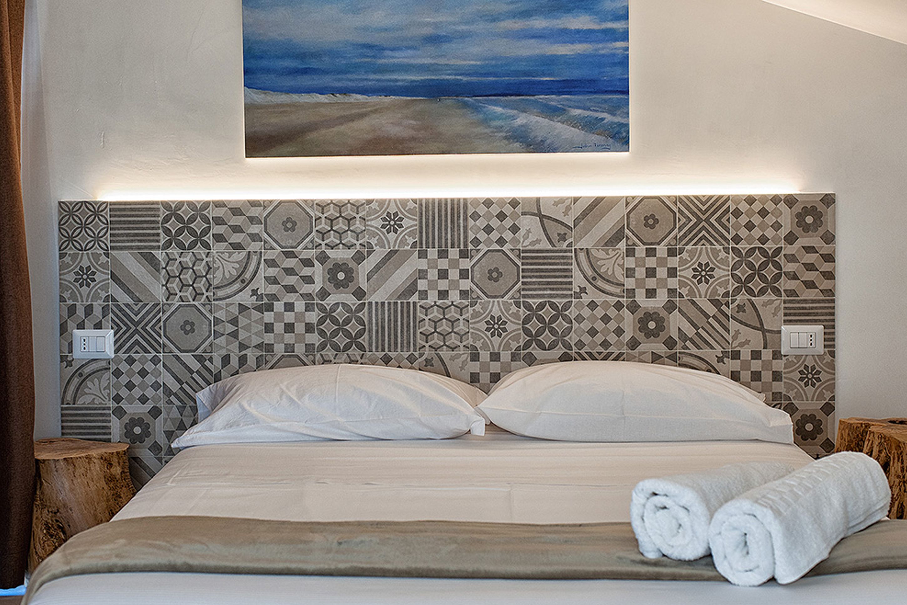 Un letto ben rifatto con lenzuola e cuscini bianchi, una testata con motivo a piastrelle geometriche e un quadro raffigurante una spiaggia sopra al letto.