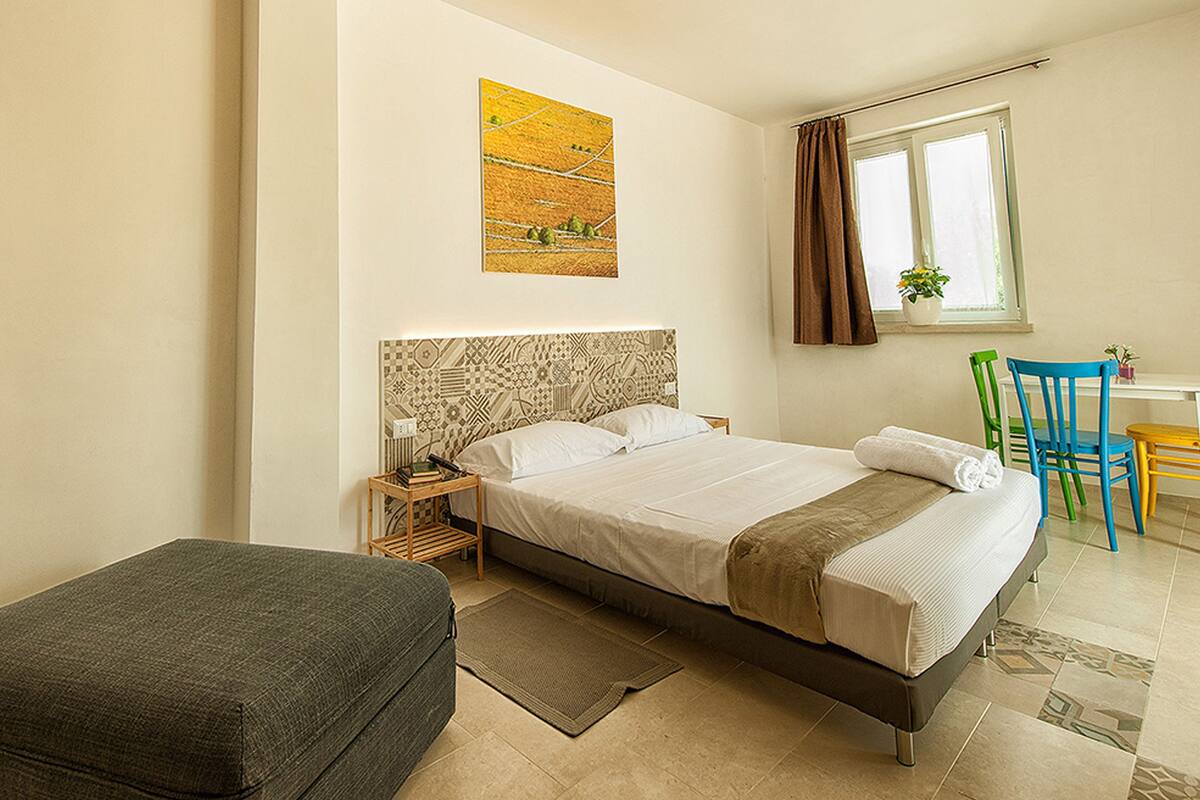 A bedroom with a bed, a nightstand, a chair, a small table, and a window with curtains.
