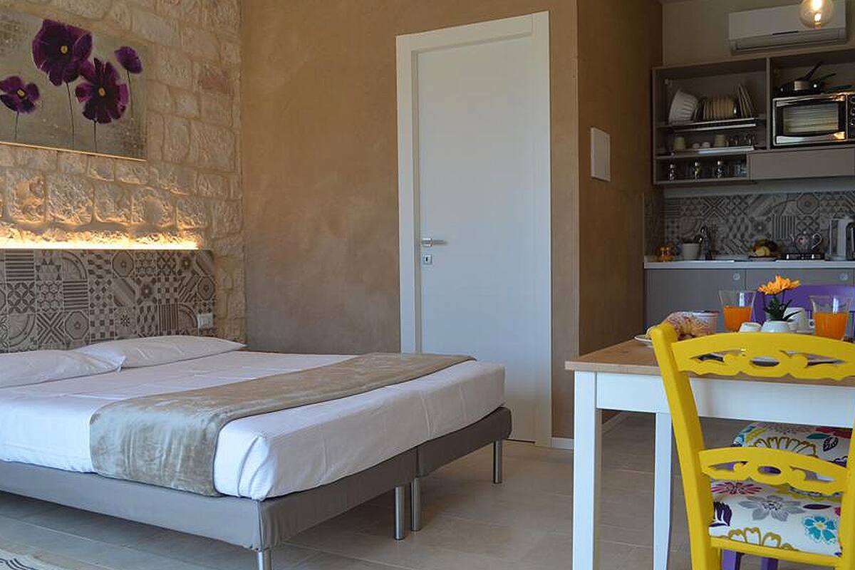 A bedroom with a bed, a white door, a small kitchenette with a microwave, and a chair with a floral cushion.