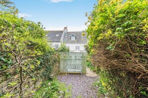 A pathway leading to a house with a white exterior and a grey roof, surrounded by dense greenery.