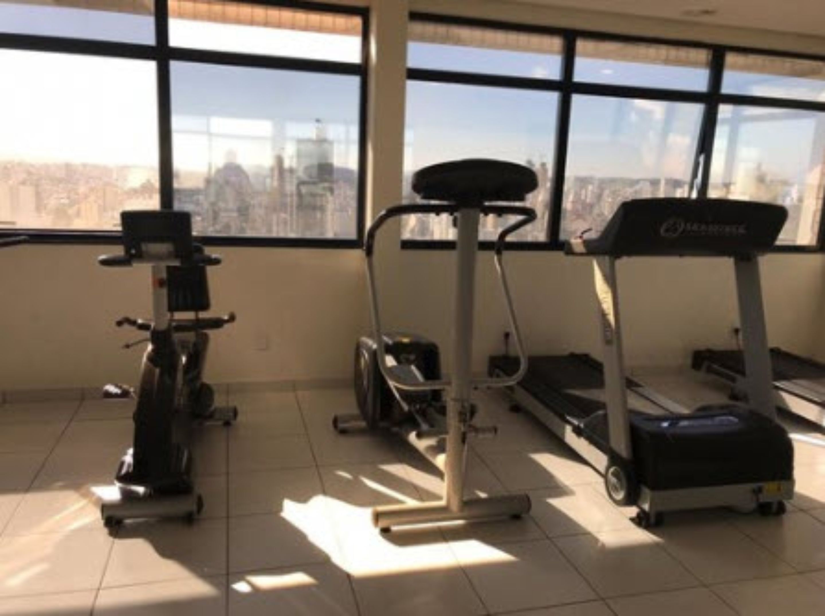 Un gimnasio con máquinas de cardio, incluyendo una cinta de correr, una elíptica y una máquina de remo, con ventanas amplias que ofrecen vistas a la ciudad.
