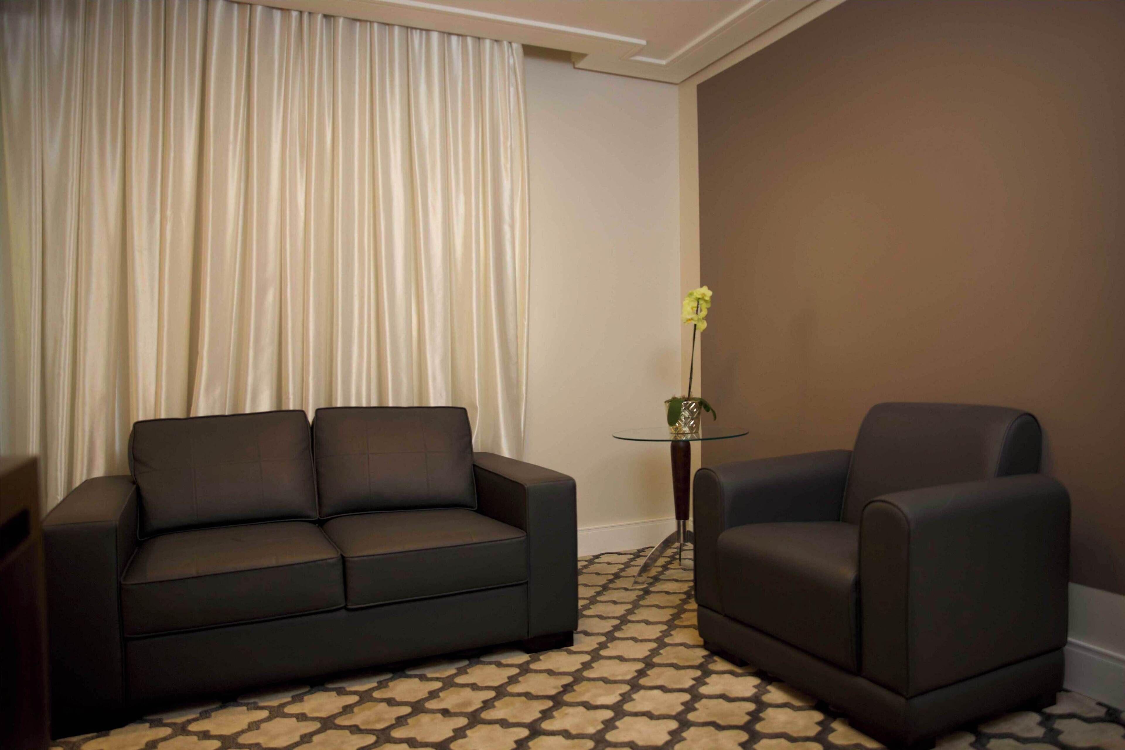 A modern living room with a dark grey sofa and armchair, a patterned carpet, a small glass table with a vase of yellow flowers, and a large window with sheer curtains.