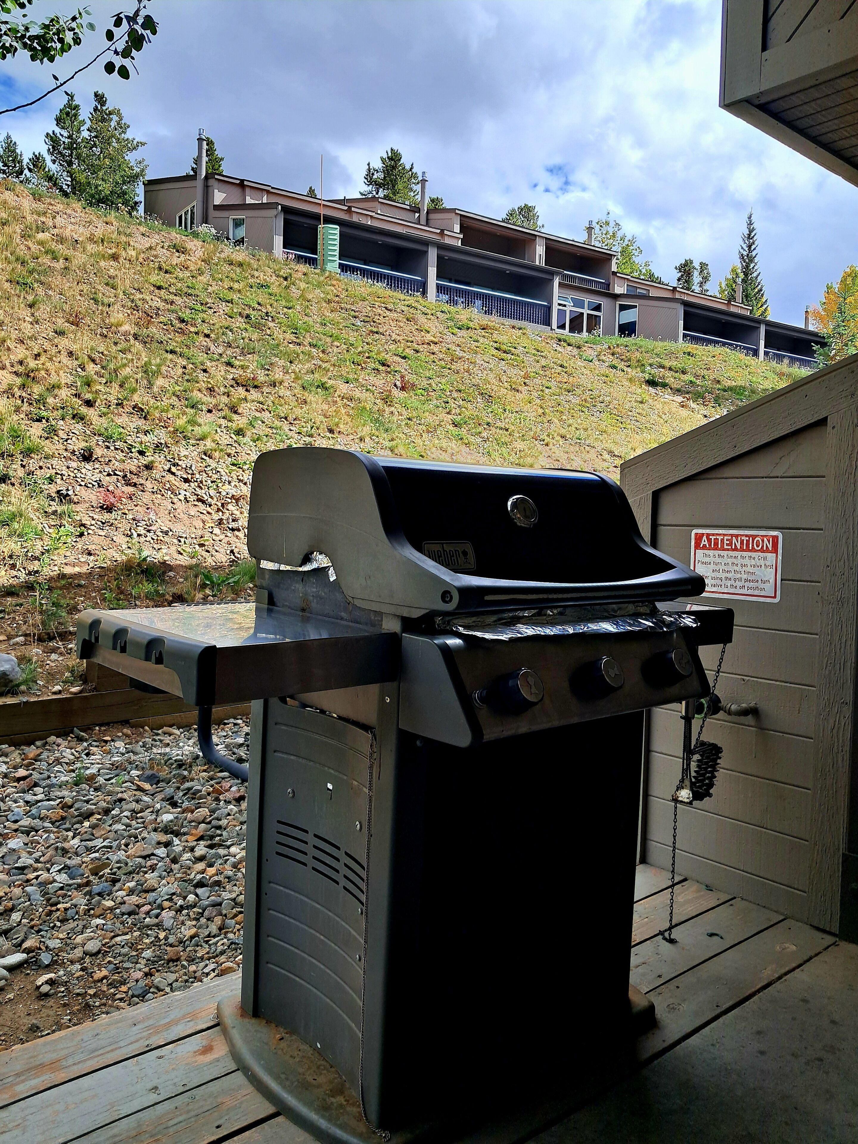 Un barbecue avec un panneau d’avertissement, une terrasse en bois et une colline avec des bâtiments en arrière-plan.