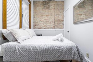 A bedroom with a bed, white bedding, a brick wall, a window with blinds, and a door. - Windy City Wanderer - Depaul, Train, Wrigley Field (Chicago)