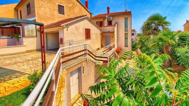 Une cour intérieure avec un escalier menant à une porte, entourée de bâtiments dotés d’un toit en tuiles et d’une cheminée rouge.