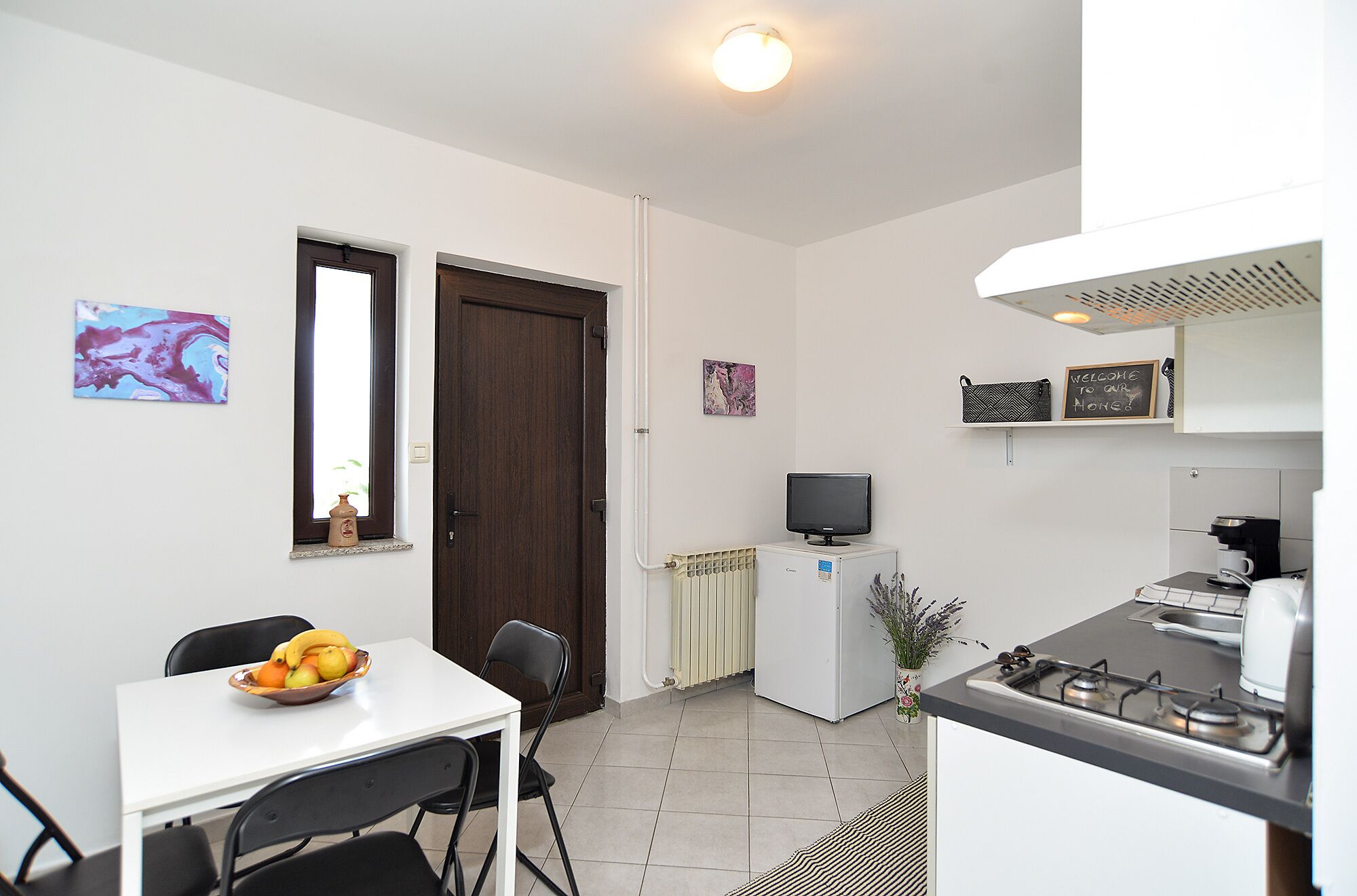 Uma cozinha compacta com área de refeições, uma televisão e um aviso que diz "Bem-vindos à nossa casa".
