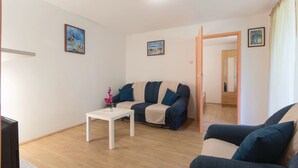 Ein Wohnzimmer mit einer Couch, einem Couchtisch und einem Fernseher. An den Wänden hängen Bilder und eine Tür führt in einen anderen Raum.