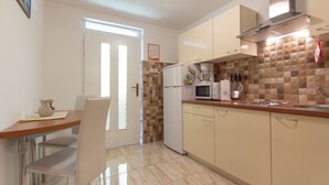 Eine kleine Küche mit Kühlschrank, Mikrowelle und Spüle. Es gibt einen Esstisch mit Stühlen und ein Fenster mit Jalousien.