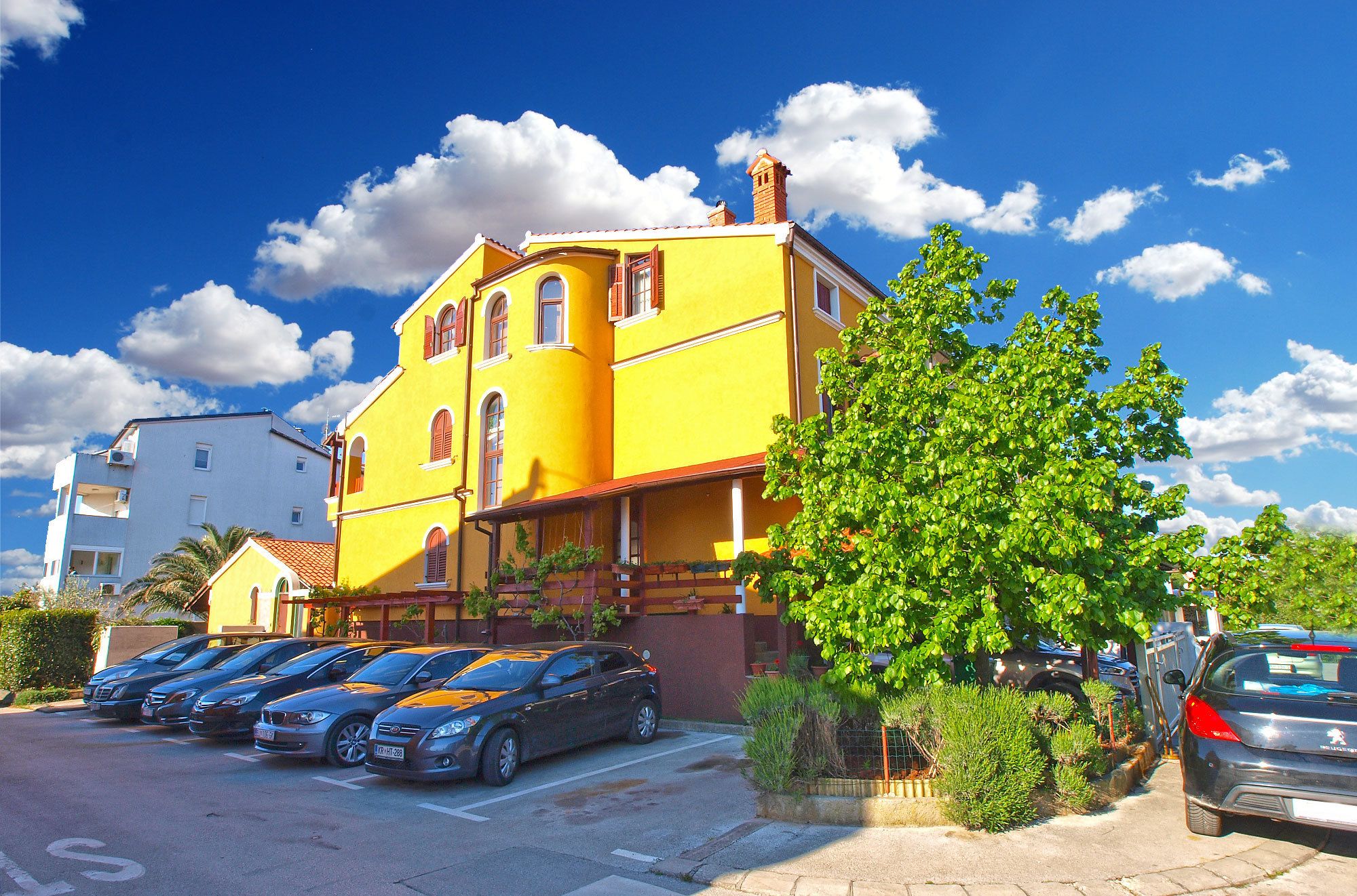 Un edificio amarillo con techo rojo, coches aparcados y un árbol en primer plano.