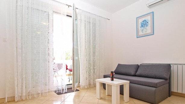 A living room with a grey sofa, a small white table, and a glass door leading to an outdoor area. - Apartment 1406 (Pula)
