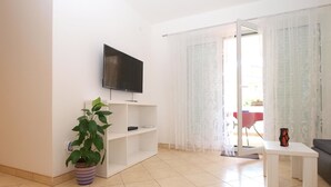 A modern living room with a flat-screen TV mounted on the wall, a white shelving unit, a potted plant, and a sliding glass door leading to an outdoor area. - Apartment 1406 (Pula)