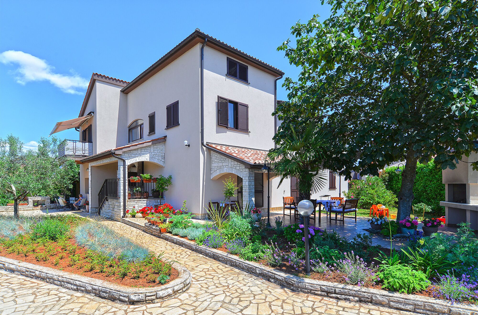 A two-story building with a balcony, a stone pathway, and a garden with various plants and flowers.
