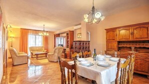 Un coin repas avec une table ronde dressée pour six personnes, entourée de chaises en bois. Un lustre est suspendu au-dessus de la table, et la pièce dégage une atmosphère chaleureuse et accueillante grâce à des meubles en bois et à une grande fenêtre agrémentée de rideaux.